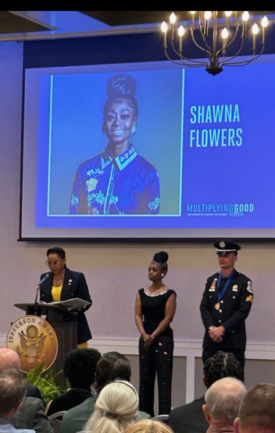 Awards ceremony with speaker, two honorees, and screen displaying "Shawna Flowers."